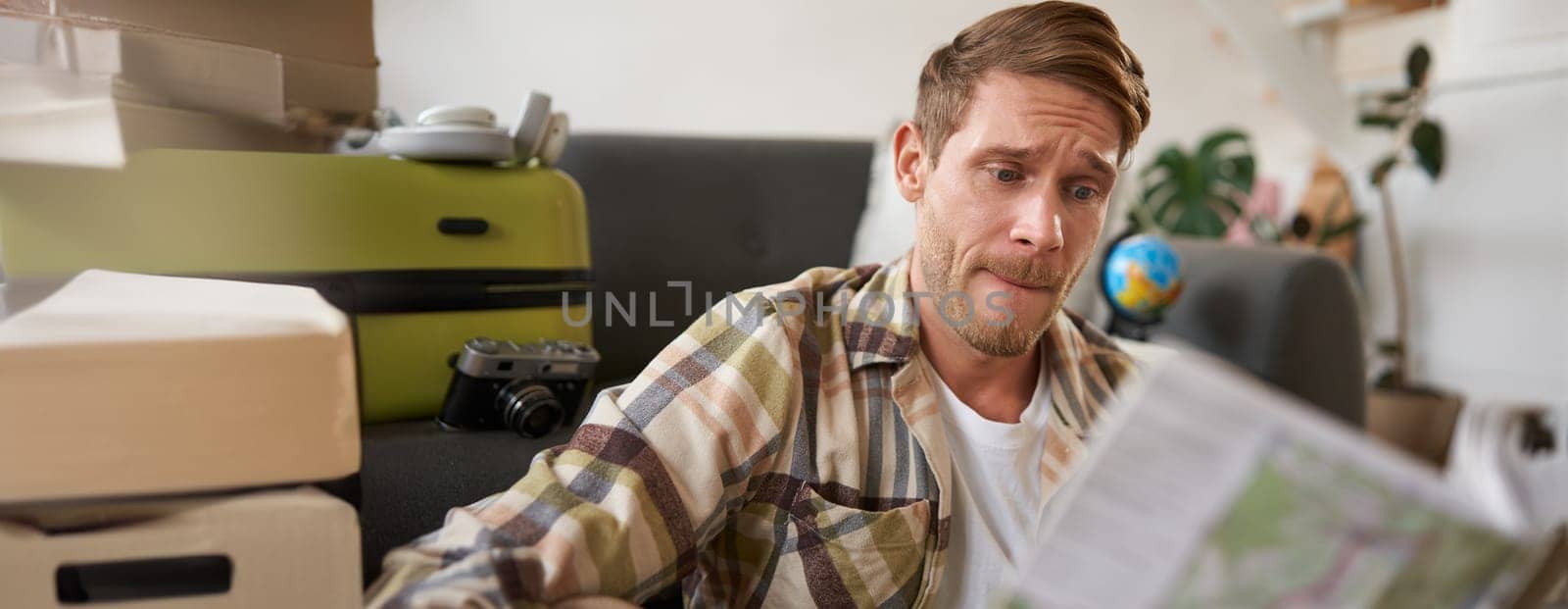 Portrait of man looking confused at travel map, choosing route, going on holiday, preparing for vacation, sitting in living room with suitcase.