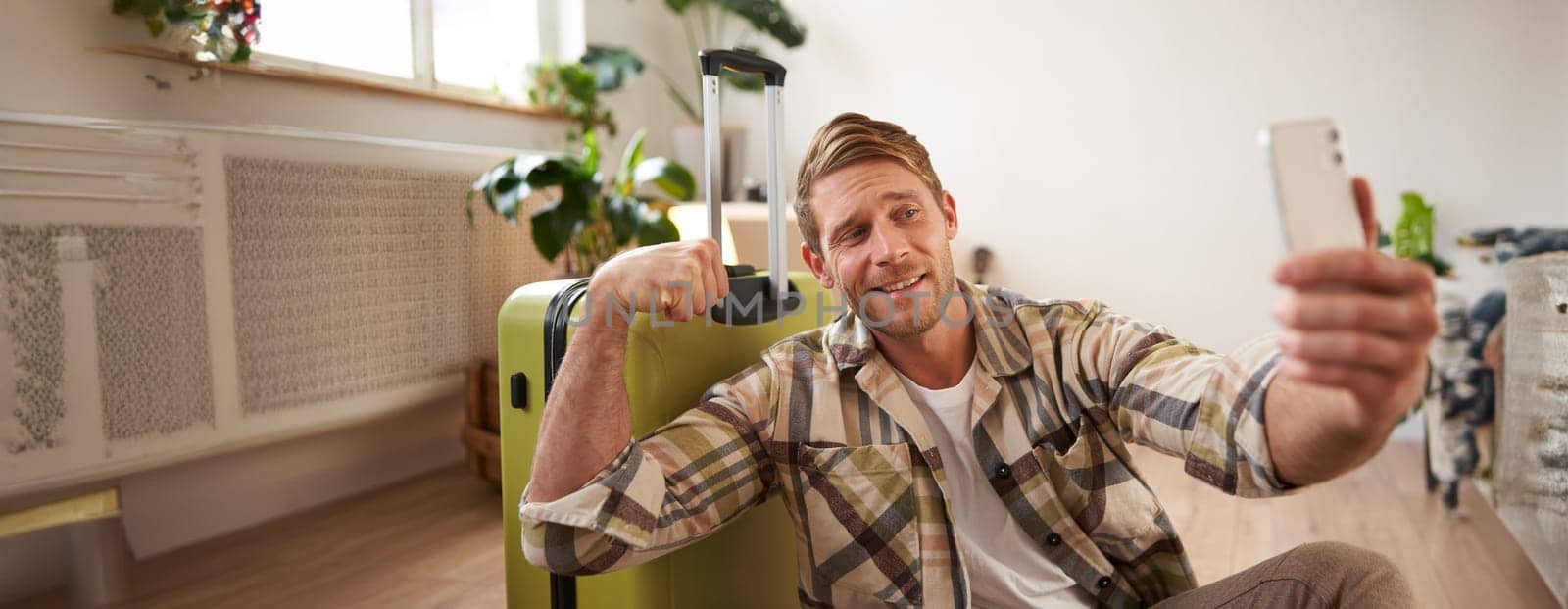 Image of handsome smiling young man going on holiday, posting online about vacation, taking selfie on smartphone with suitcase, shows muscles.