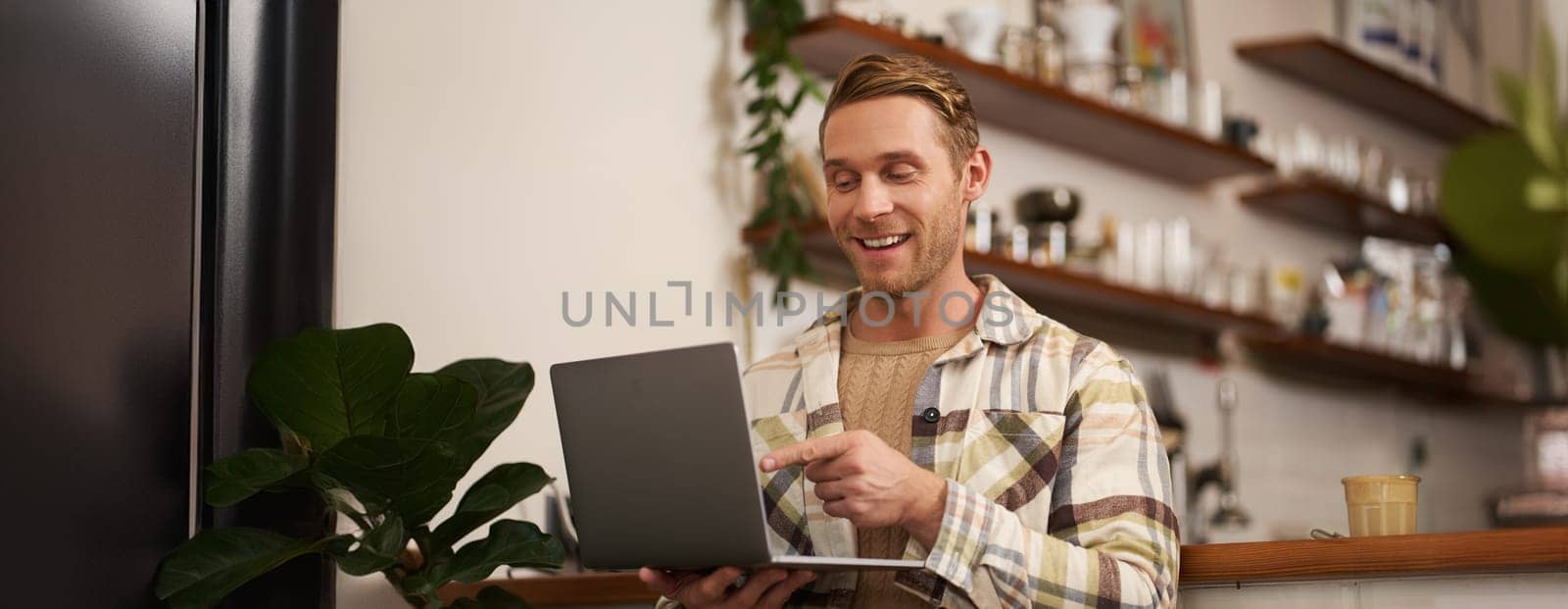 Image of young man in coffee shop, connects to video call, talking online, chats with friends via laptop and smiles.