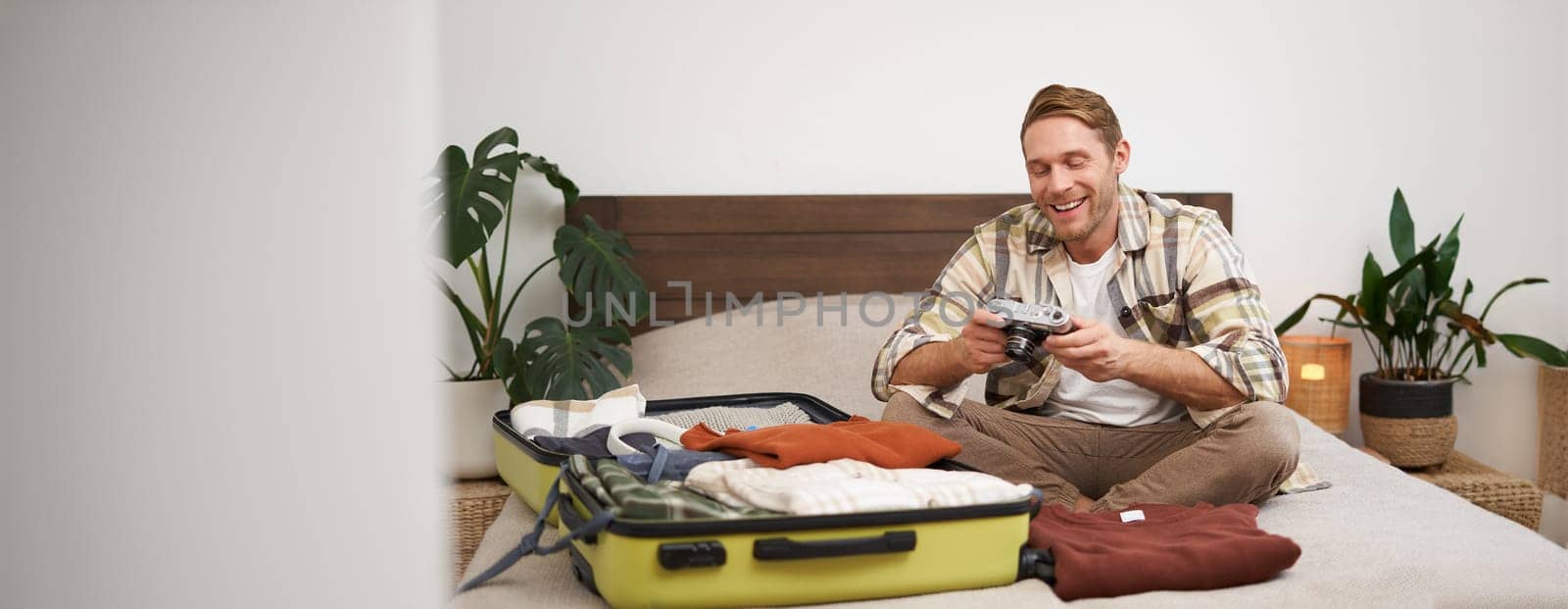 Portrait of happy, smiling young man sitting on bed, unpacking his suitcase after a holiday, tourist looking at his photo album on camera.