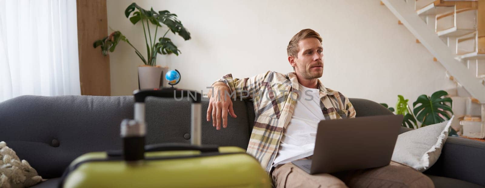 Portrait of happy young man, tourist booking his holiday with travel agency, looking at laptop, packed suitcase to go on vacation.
