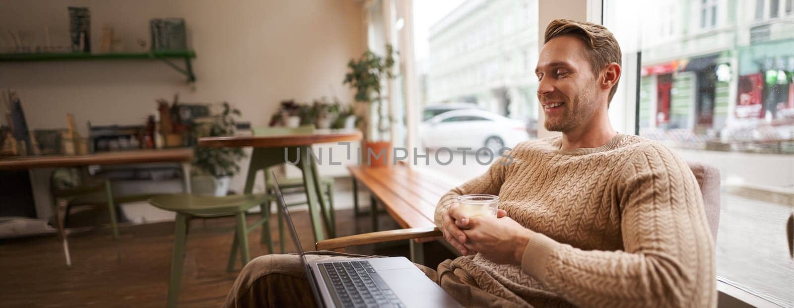 Portrait of handsome young man sits in cafe, drinks coffee and watches video on laptop, looking at screen with happy smile, relaxing in co-working space.