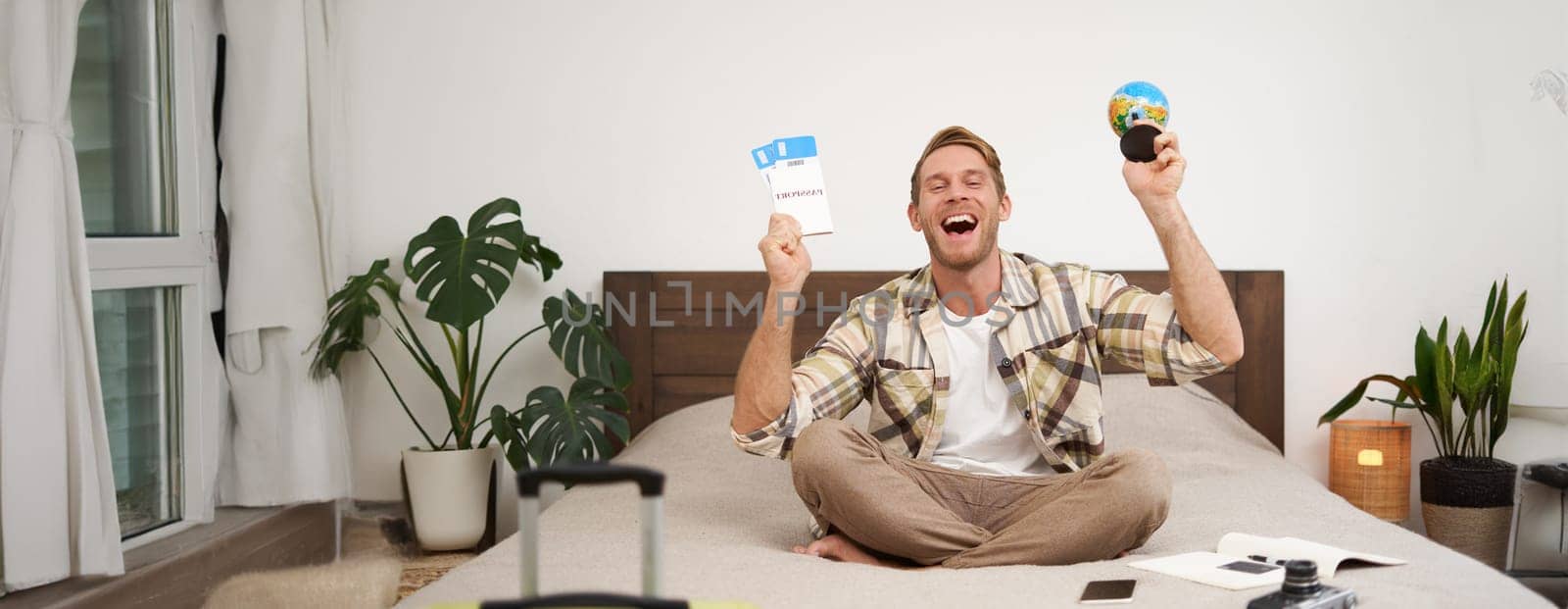 Portrait of cheerful, happy tourist, man sitting with plane tickets and a globe, packed suitcase for travelling around the world, going on vacation, excited about his holiday.