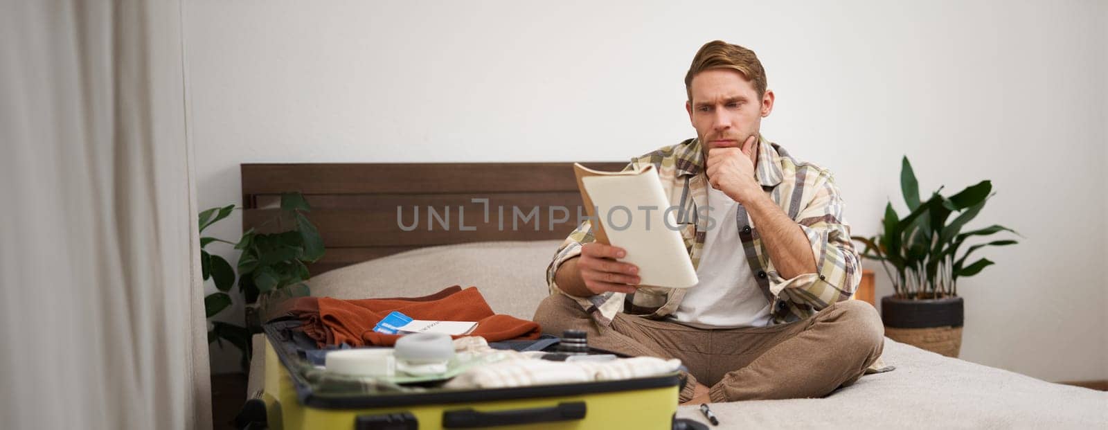Portrait of man with suitcase, sits in room and looking serious at his notes, packing for holiday, checking list of items in notebook. Copy space