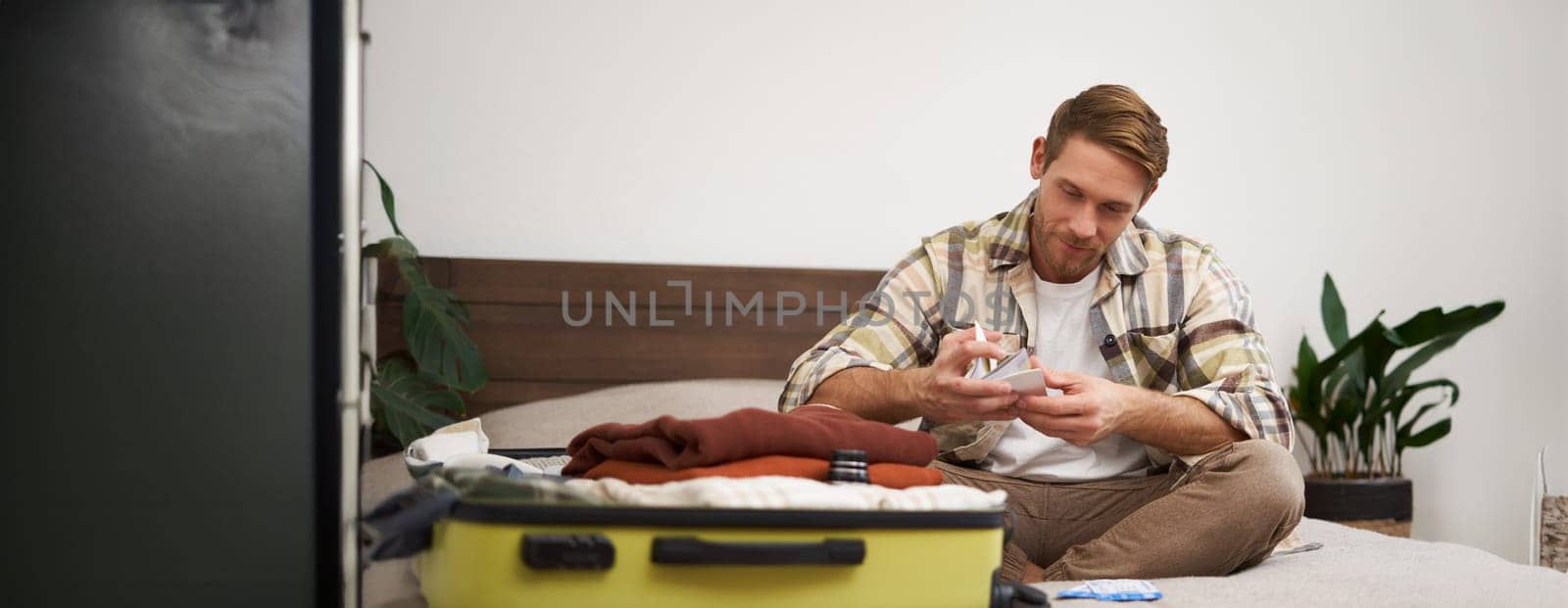 Portrait of young man with suitcase, holding his plane tickets, preparing to go on holiday, packing things for vacation, tourist puts clothes in bag.