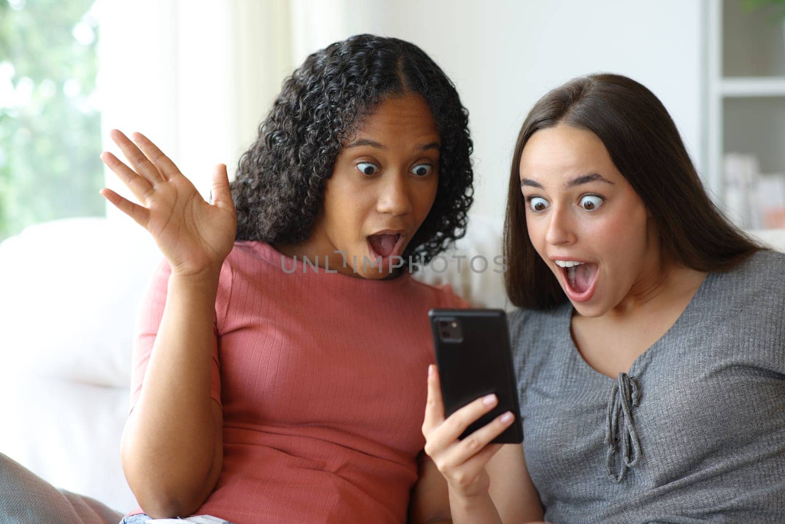 Amazed interracial friends checking cell phone content sitting on a couch at home