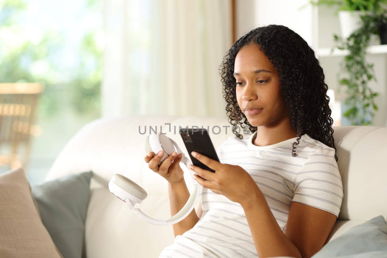Black woman pairing wireless headphone with smart phone sitting on a couch at home