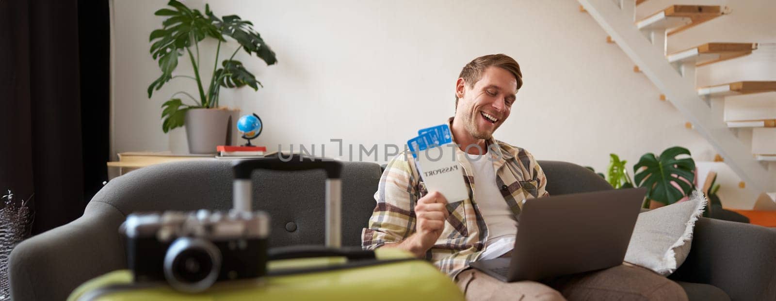 Portrait of happy tourist, man looking excited at laptop screen, holding flight tickets, going on vacation, excited about holiday, sits with suitcase in living room.