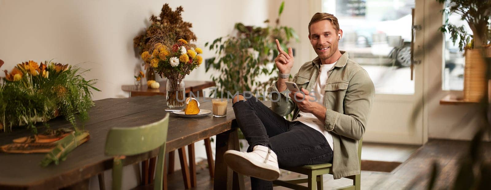 Portrait of happy handsome young man, sitting in cafe with mobile phone, wearing wireless headphones, listening to music or podcast while relaxing in coffee shop.