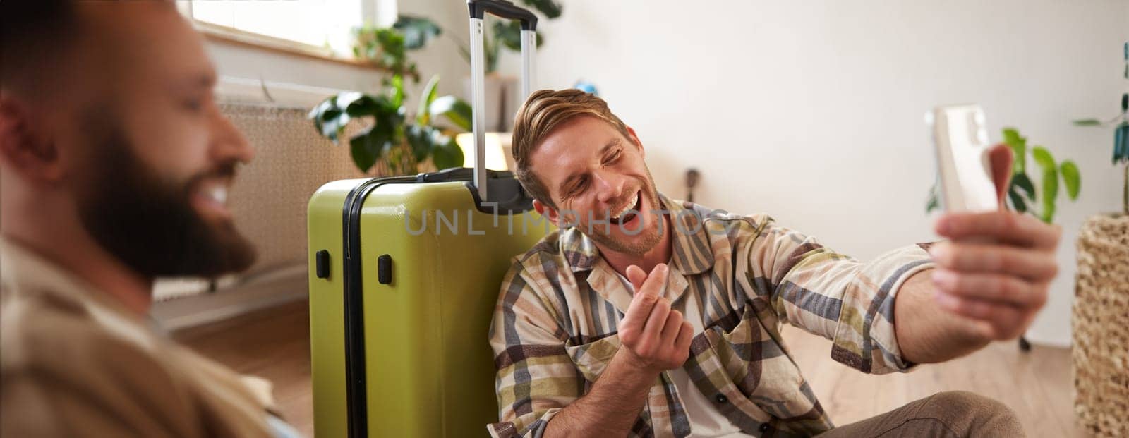 Smiling young man with suitcase, takes selfies on smartphone, blogger posts about his holiday tour on social media, using mobile phone. Tourism and travelling people concept