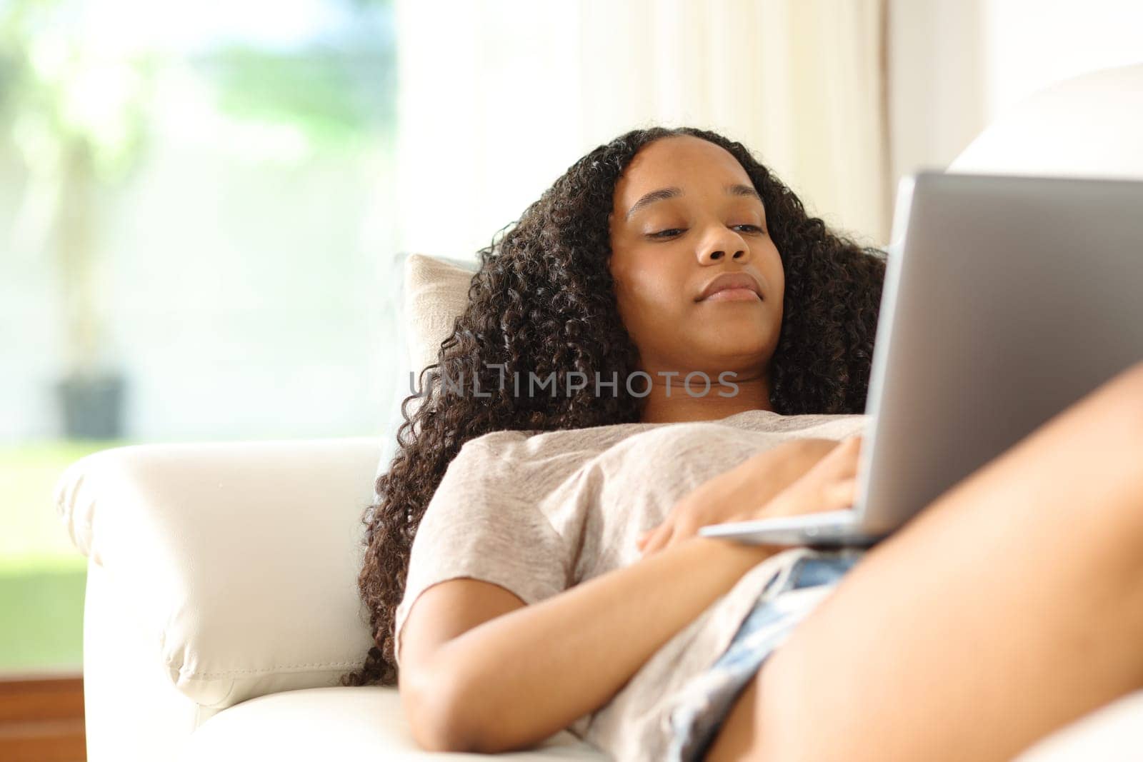 Black woman at home checking laptop content lying on sofa