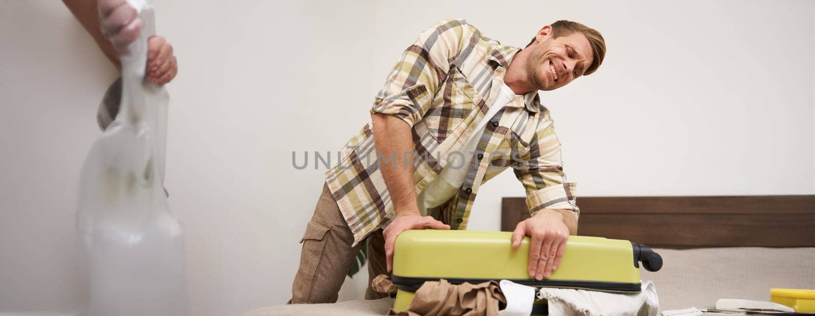 Image of young man, tourist struggling to close his suitcase full of clothes, pushing on bag, trying to press items inside luggage to shut the zipper, packing for travelling abroad, going on holiday,