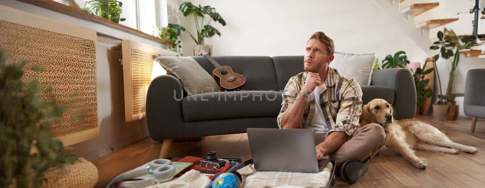 Portrait of young man, traveller going on holiday with a dog, packing his suitcase, sitting with laptop on his luggage, thinking, making online booking.