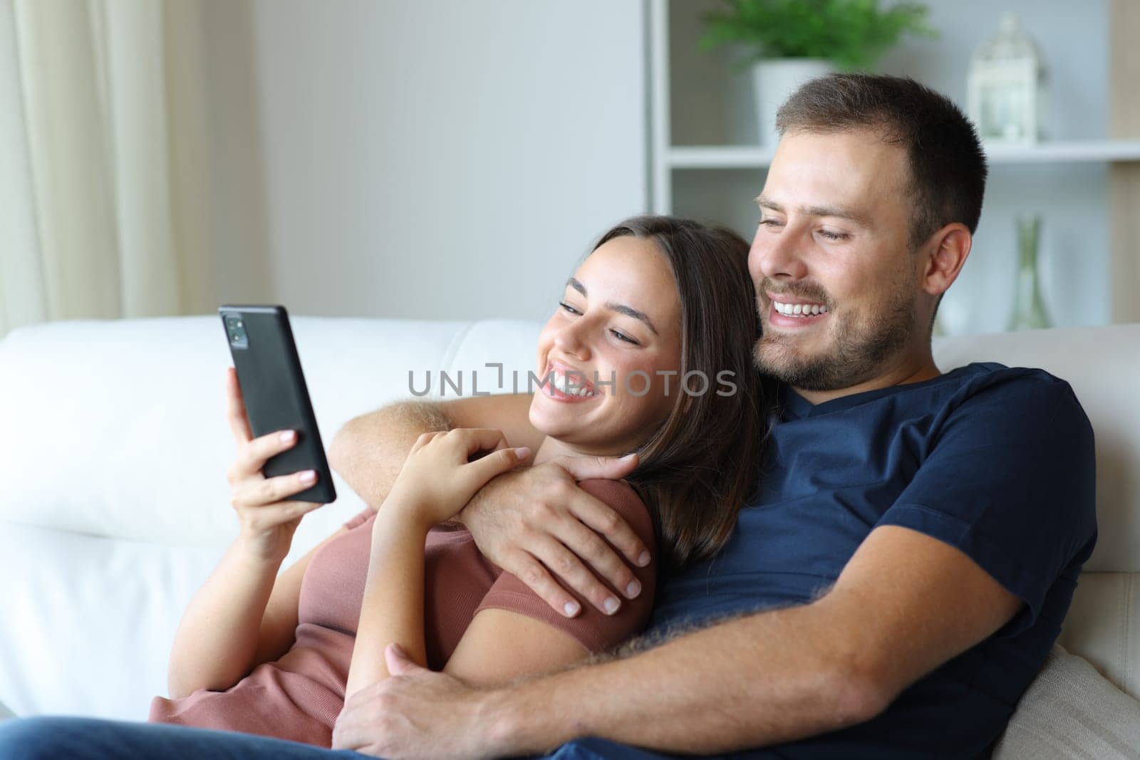 Happy couple using smart phone hugging sitting on a sofa at home
