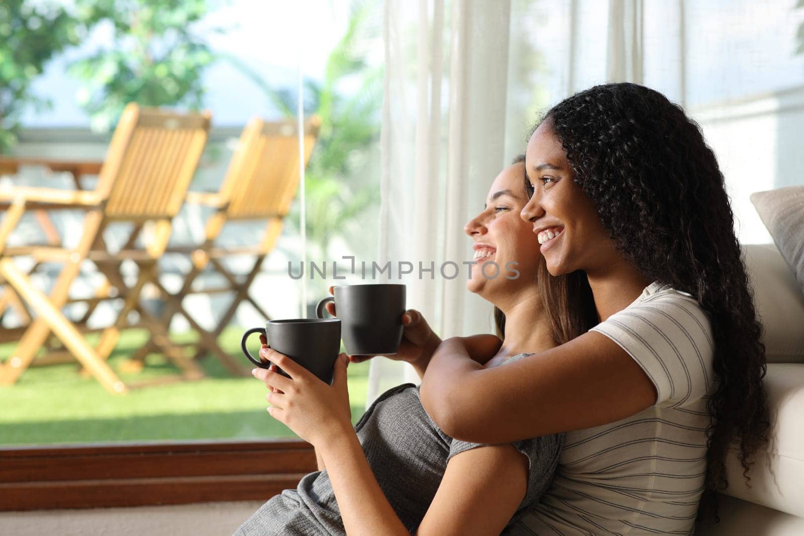 Happy interracial lesbian couple drinking coffee and contemplating through a window at home