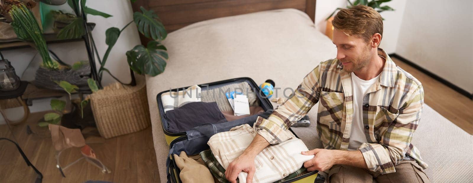 Portrait of handsome young man going on holiday, guy prepares for a trip journey, packs his suitcase, puts clothes in luggage, gets ready for adventure.