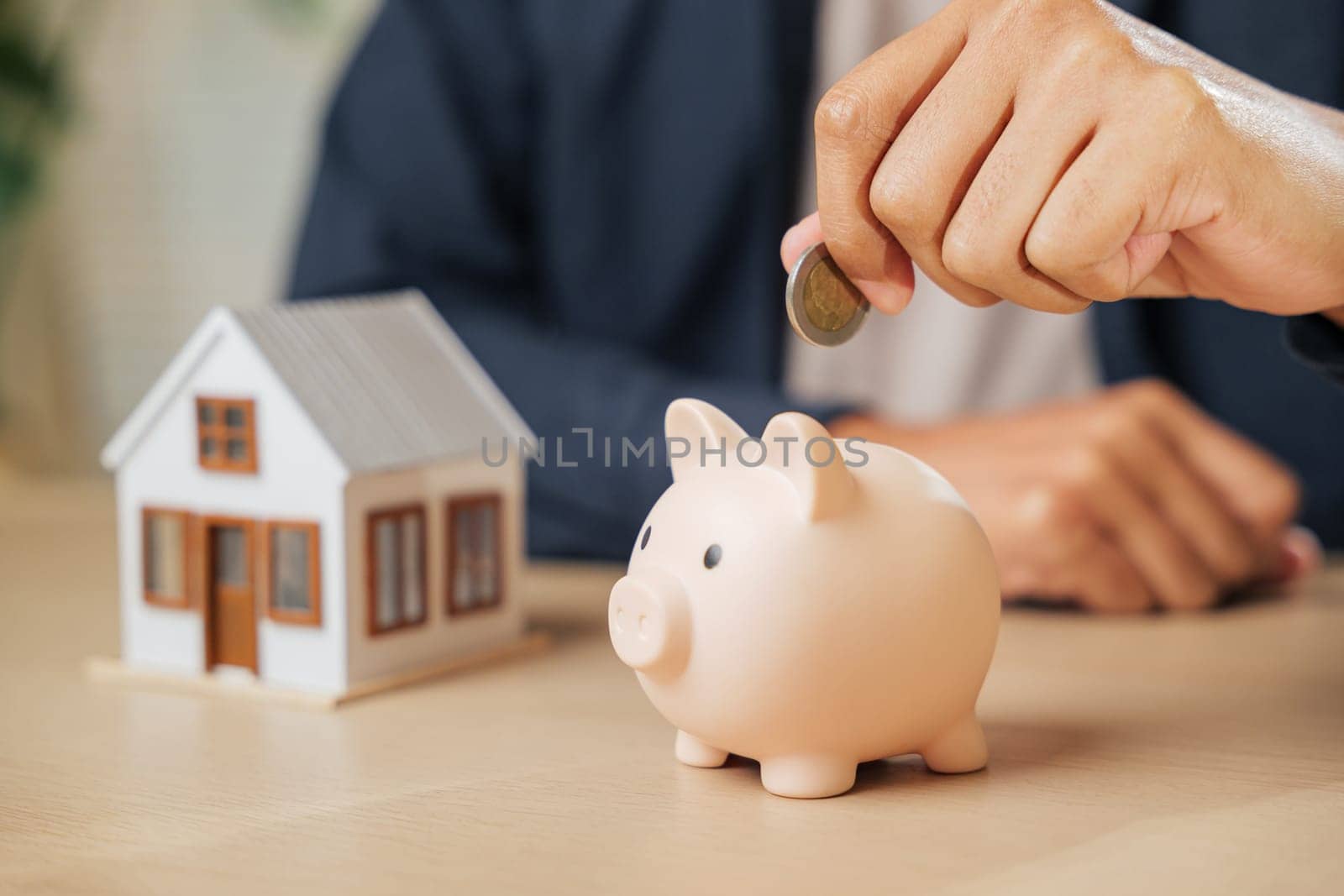 A businessman hand saving money by placing a coin into a piggy bank next to a house model, representing investment, financial stability, property goals, and a savings-focused approach to home buying.