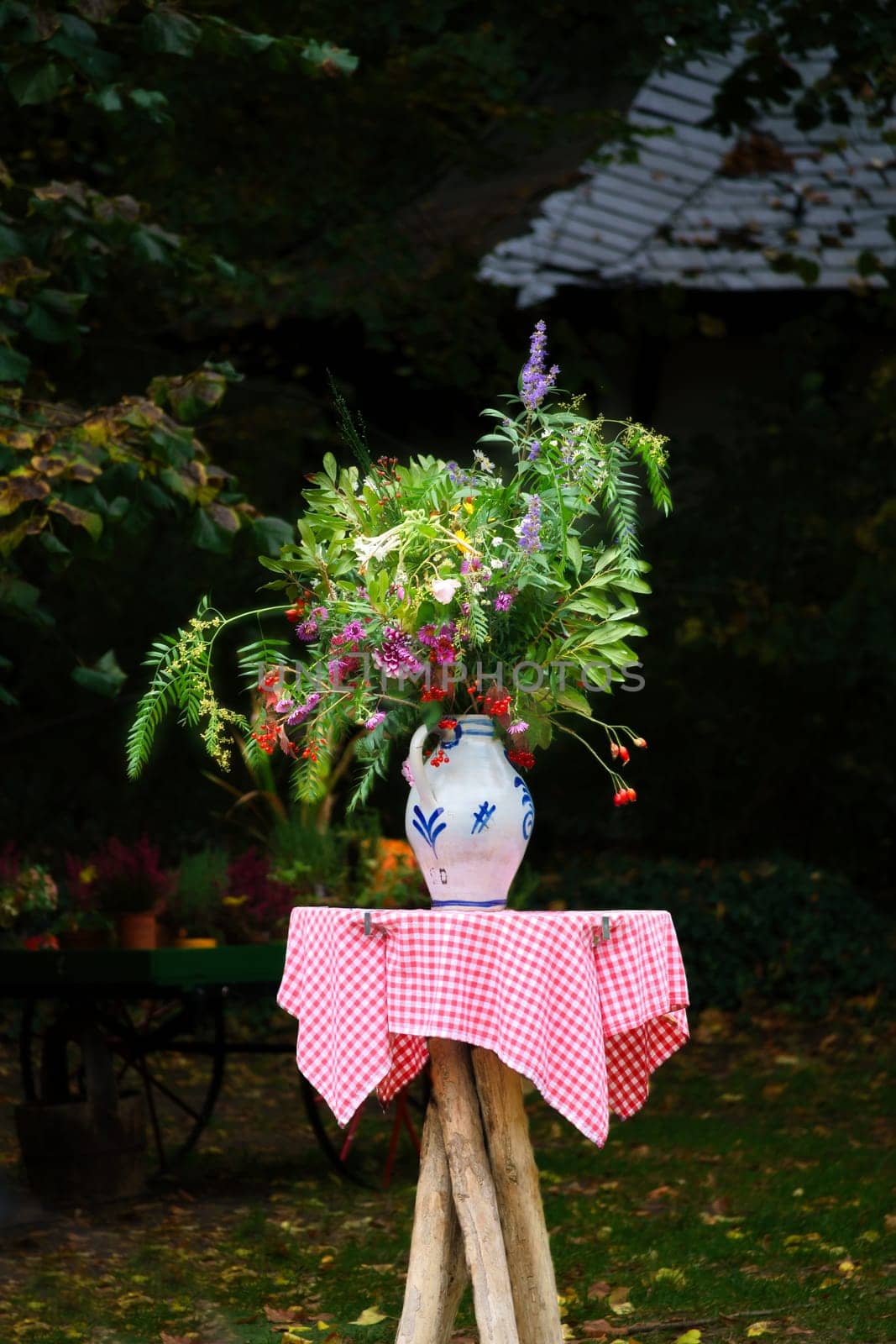 A bouquet of flowers in a vase on a table with a red and white checkered cloth, in the background a garden with trees and plants. Ideal for nature and garden lovers.