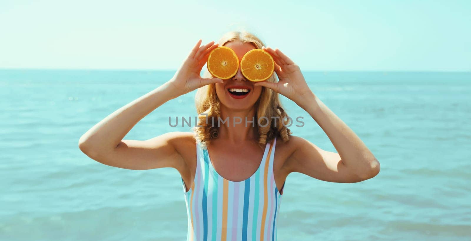 Summer vacation, portrait of happy smiling young woman, laughing, holding oranges fruits, having fun, wearing a swimsuit on the beach on sea coast background