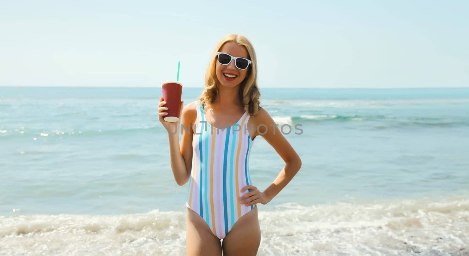 Summer vacation, beautiful happy slim cheerful woman standing in swimsuit with sunglasses having fun, holds cup of juice on the beach on sunny day on sea coast with blue sky background
