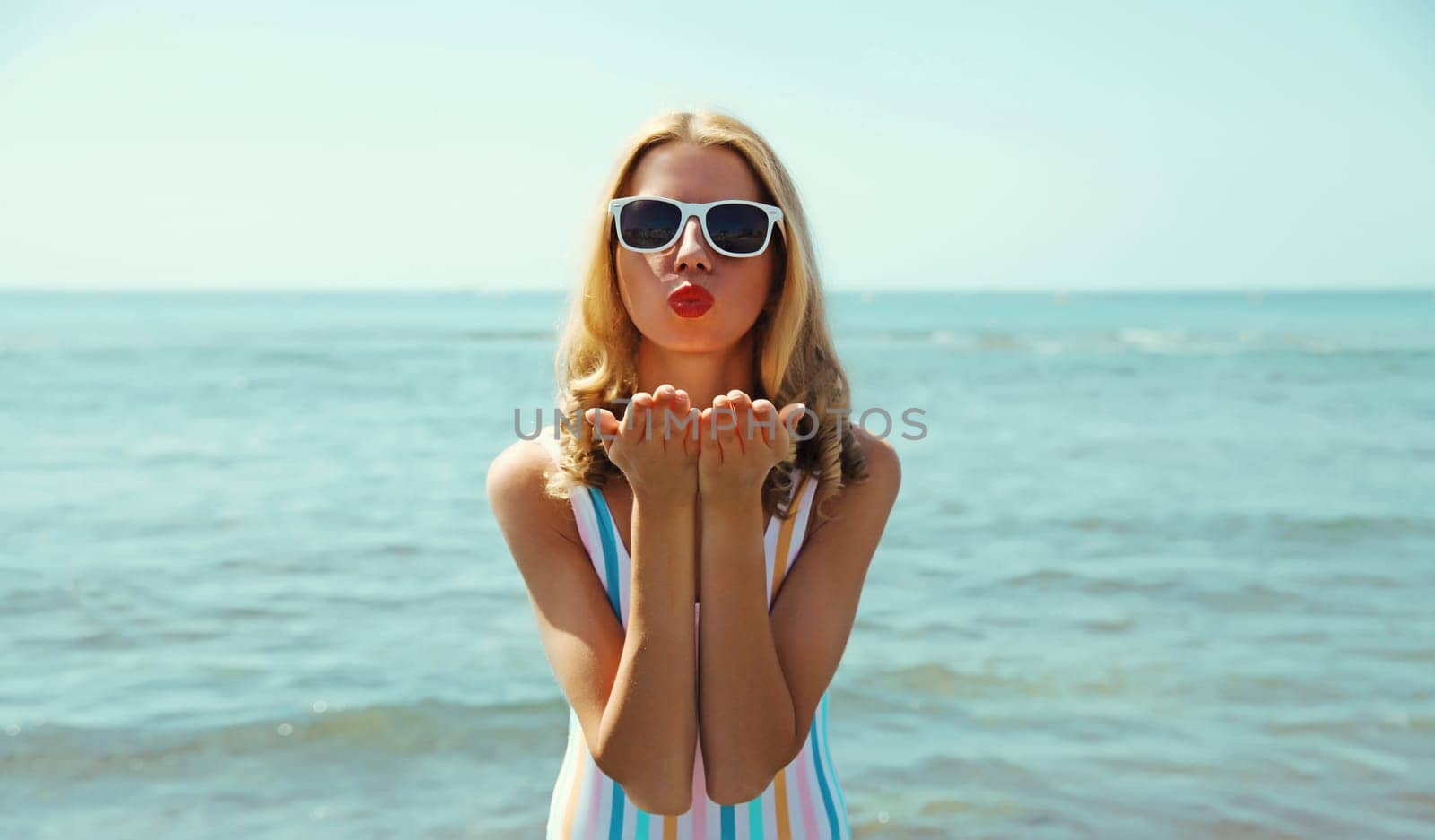 Summer vacation, beautiful happy lovely woman blowing a kiss in swimsuit, sunglasses on the beach on sea coast background