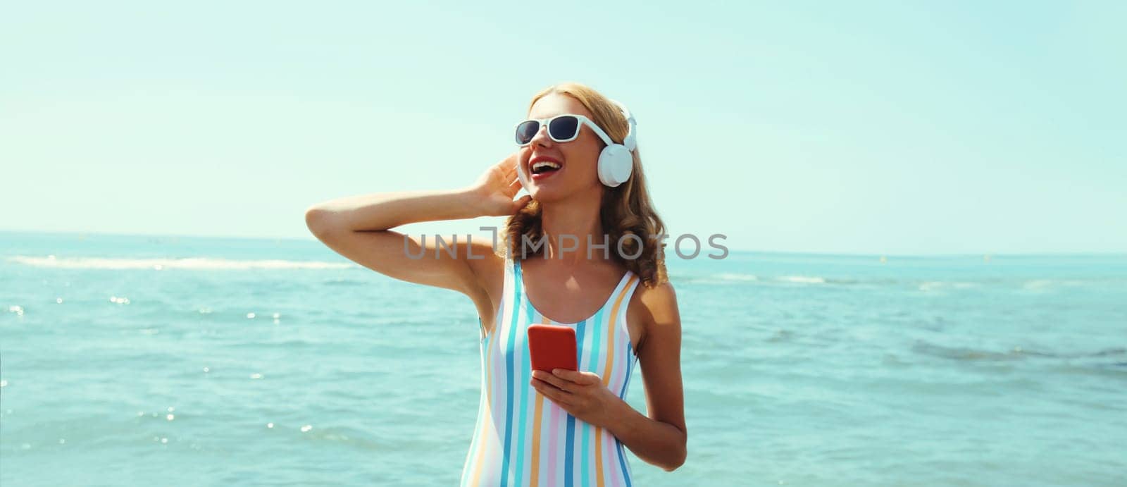 Summer vacation of happy young woman smiles enjoying listening to music with headphones, holding phone, relaxed cheerful girl wear a sunglasses on beach at sea coast with blue sky on a sunny day