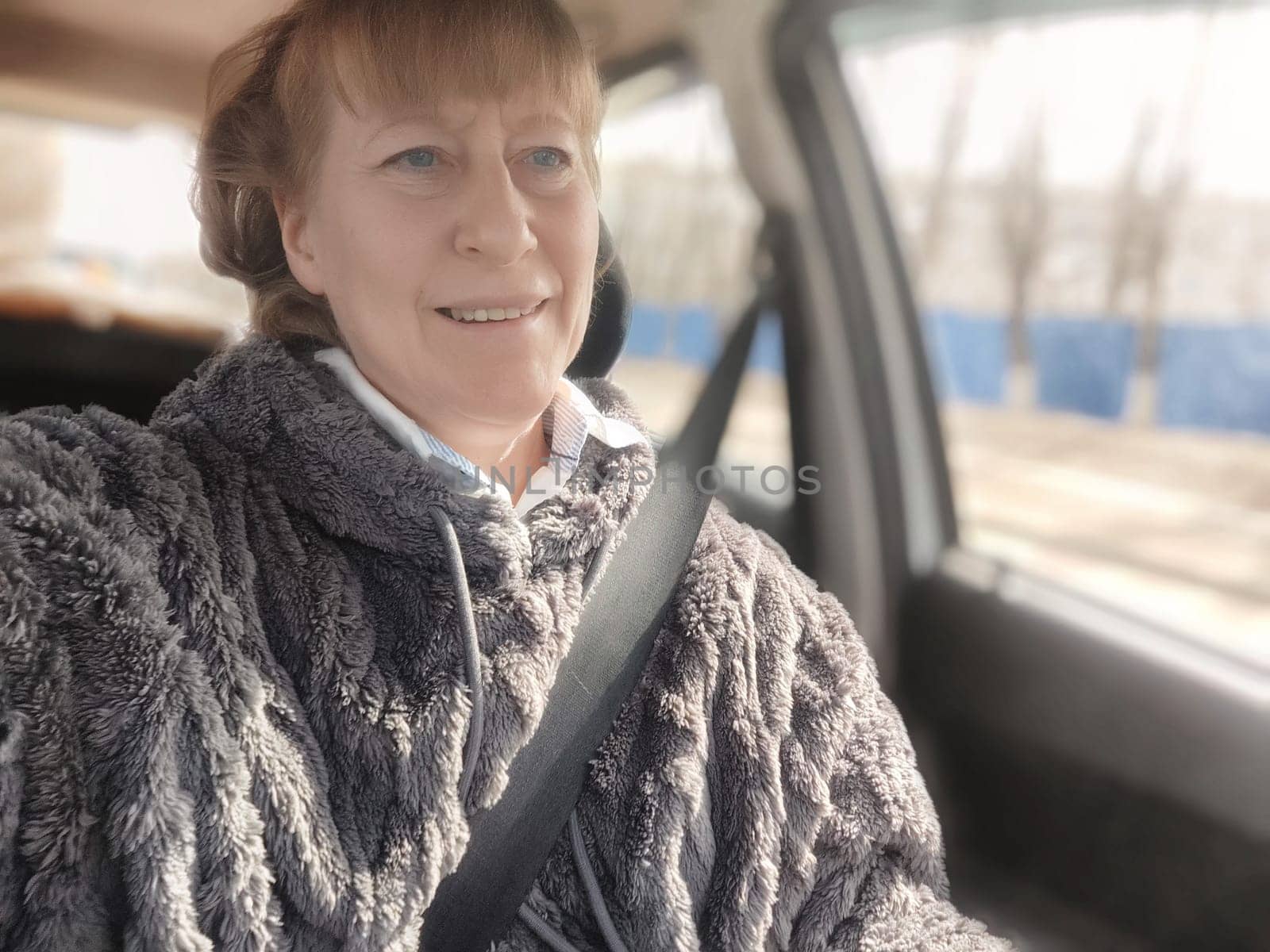 Blonde woman enjoys a cozy moment in her vehicle, capturing a selfie while driving and connecting with her blogging audience on a bright day.