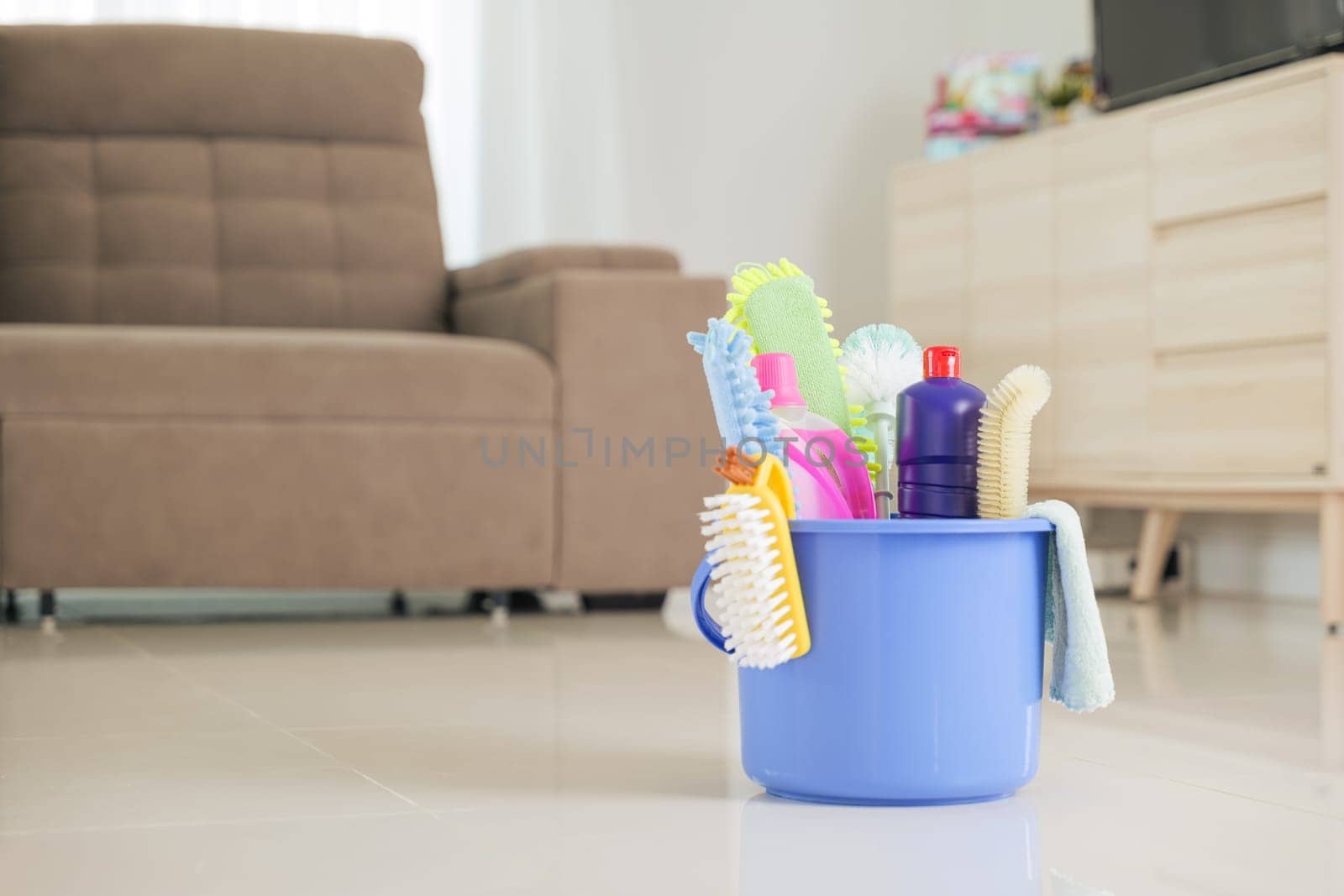 Cleaning tools and supplies neatly stored in blue bucket. Detergent bottles, sponges, and brushes sit on floor in living room, making housework and daily cleaning tasks more convenient and organized.