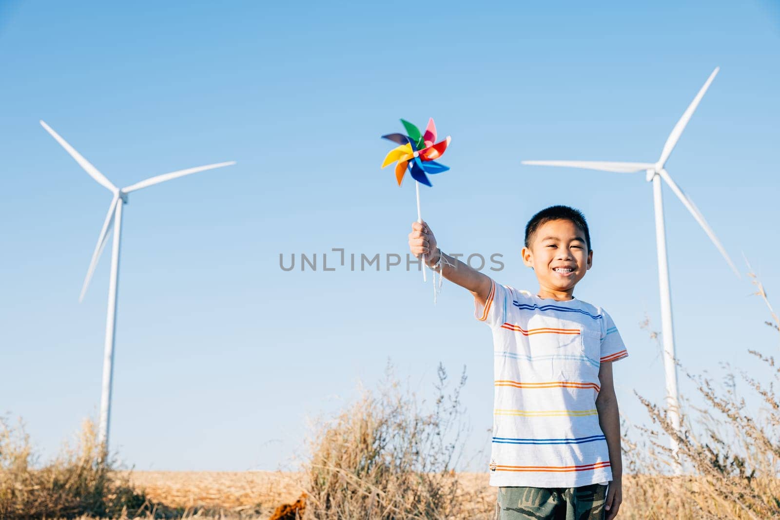 Boy's playful engagement near wind turbines holding a pinwheel toy represents fun of wind energy. Showcases clean electricity innovation in a countryside windmill landscape against a clear blue sky.