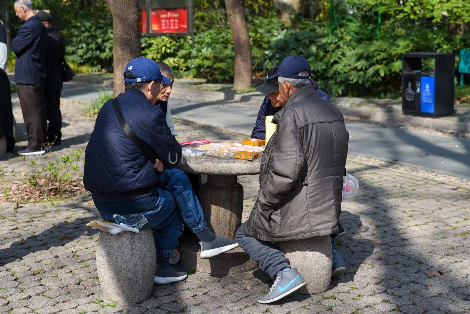 Shanghai, China – 1 April 2025: Elderly Chinese men play Xiangqi (Chinese chess) around a stone table in People's Park on a sunny day, enjoying leisure and tradition