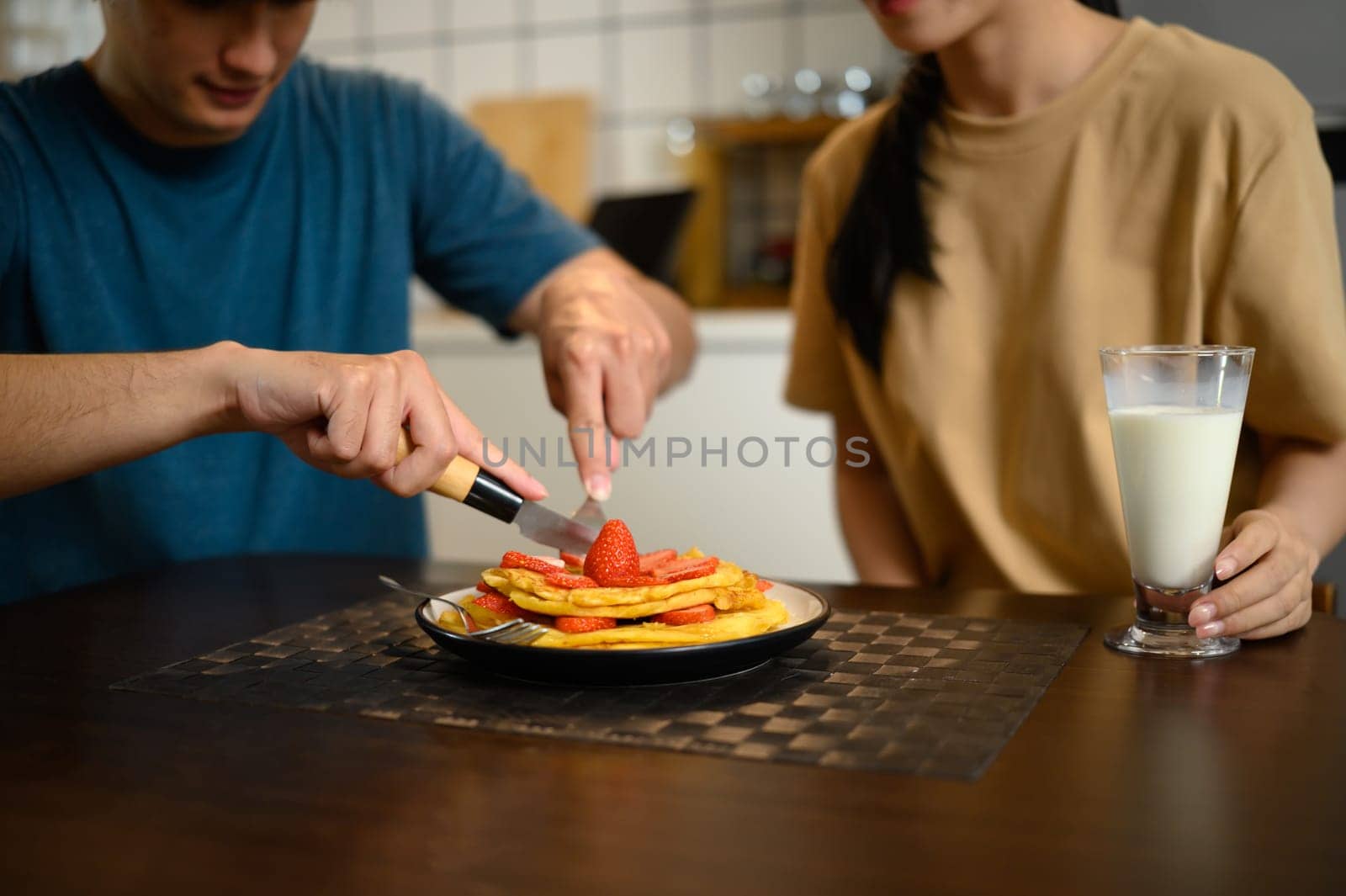 Happy young couple enjoying pancakes with strawberries and syrup in cozy home kitchen.