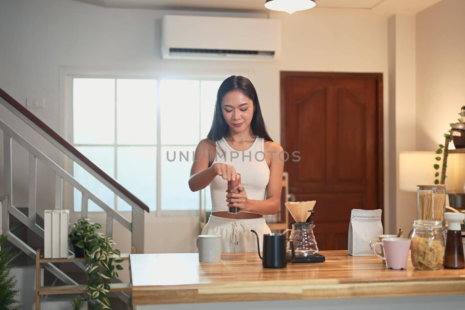 Young woman grinding coffee beans and preparing pour over coffee in a warm kitchen.