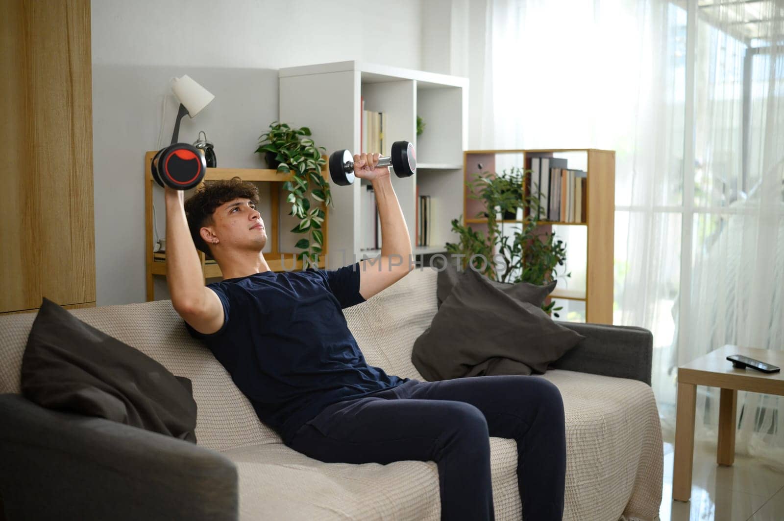 Young man lifting dumbbells on a sofa. Fitness at home concept.
