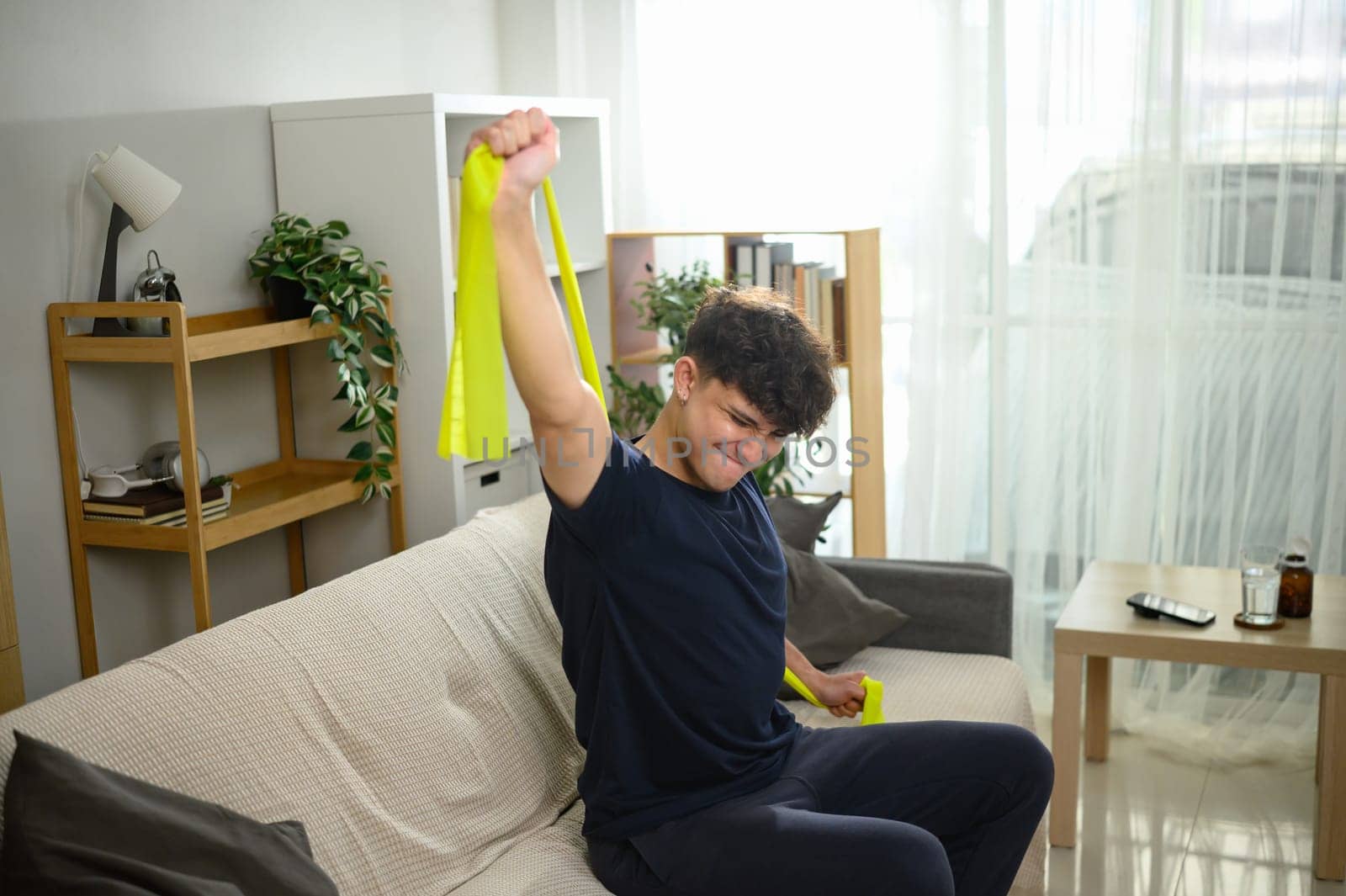 Young man exercising with yellow resistance band while sitting on sofa at home.