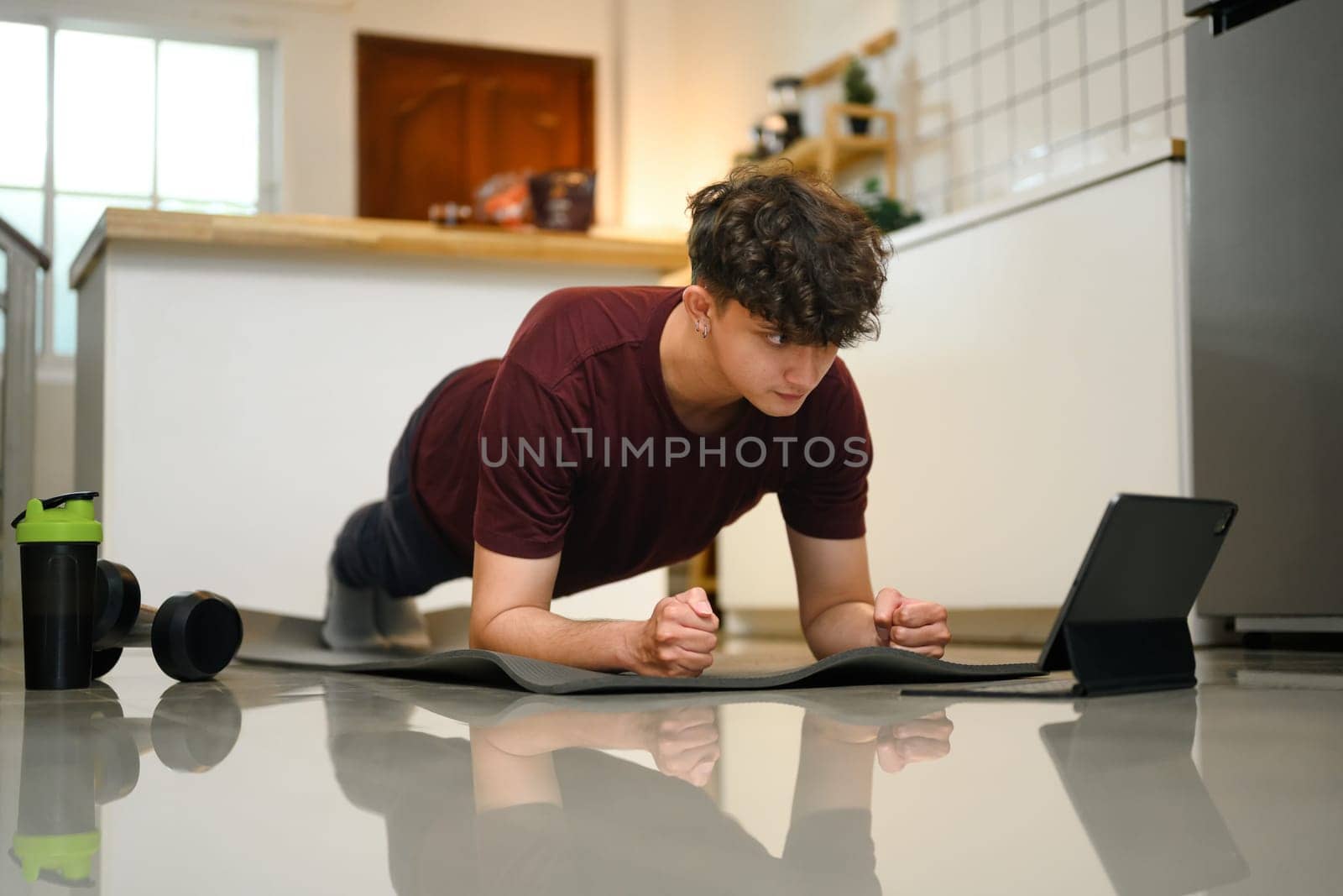 Young man doing plank exercise at home while following a workout video on a tablet.