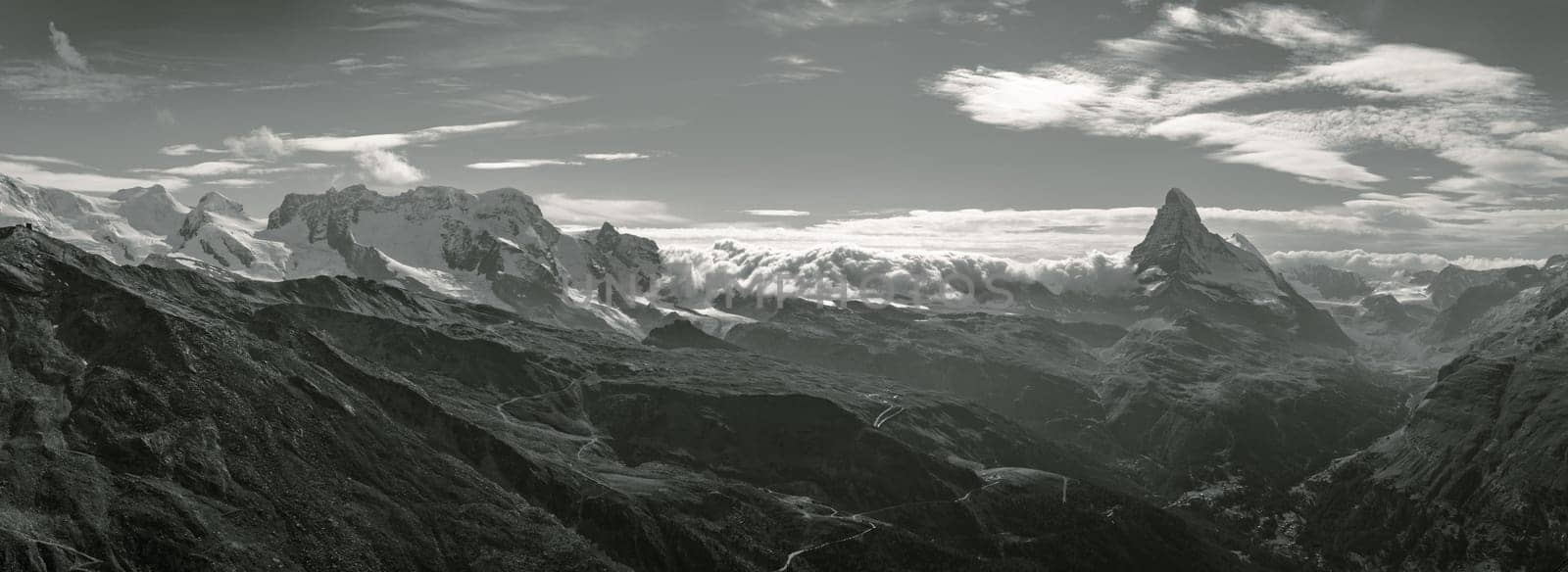 Swiss panoramic landscape photograph of the mountain range near Zermatt, with the iconic Matterhorn towering over beautiful valleys in an alpine scene. Real composite long pano photo in monochrome.