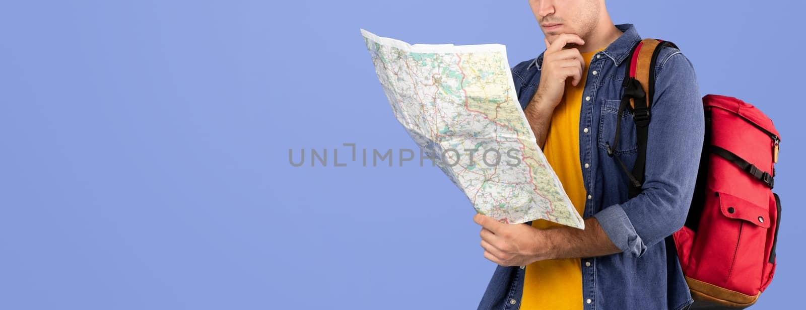 Portrait of thoughtful young hiker with rucksack looking at map over blue studio background. Pensive male tourist planning his camping trip route. Traveling and active lifestyle concept