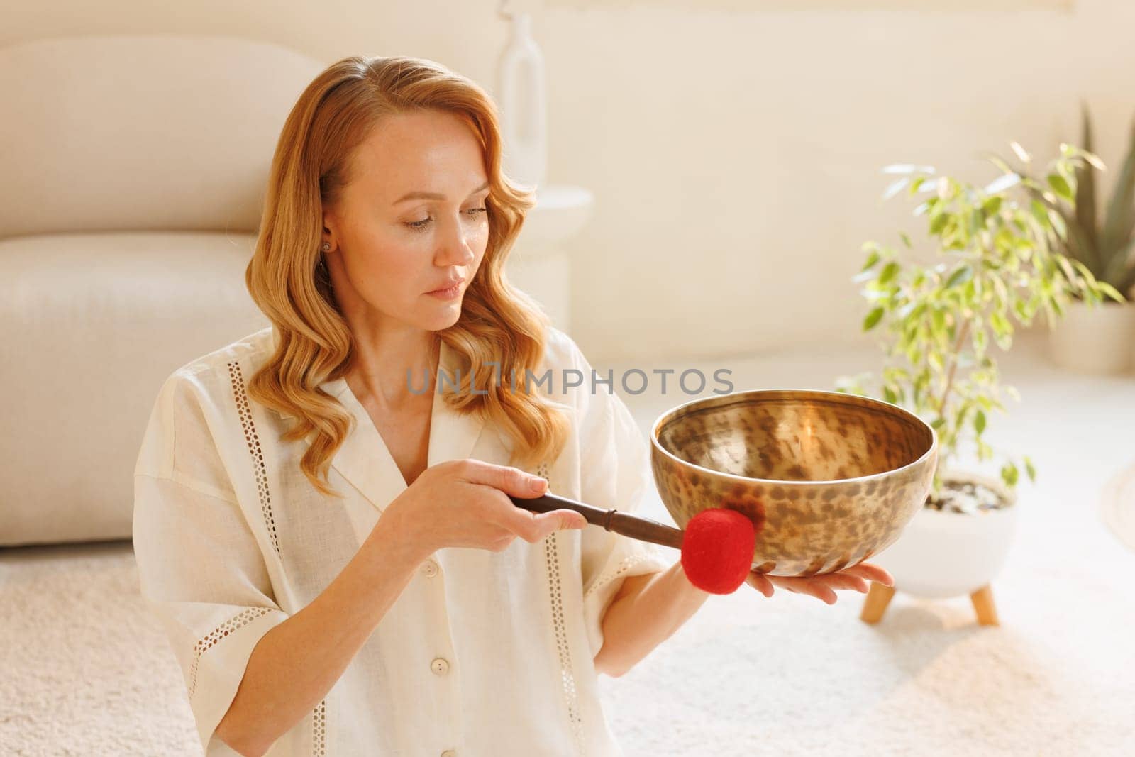 Calm woman using Tibetan singing bowl for sound therapy and meditation in a peaceful home interior with soft light.