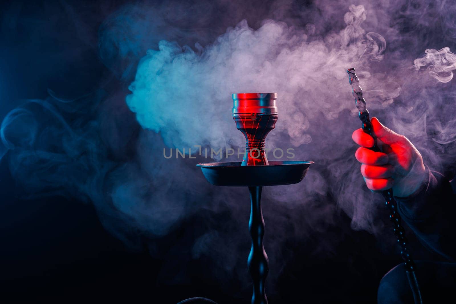 Hookah with dense smoke and vibrant red-blue lighting, hand holding pipe hose in dark moody background.