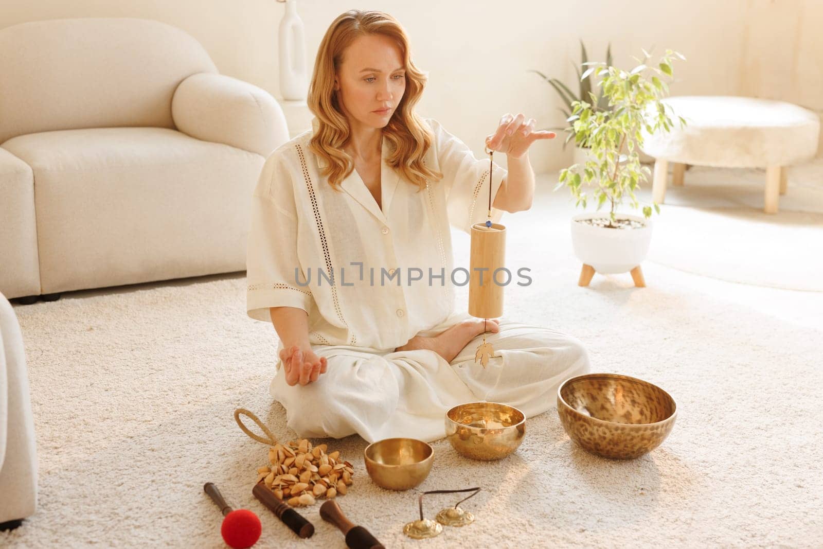 Woman meditating with a bamboo chime, surrounded by sound healing tools, in bright peaceful home interior.