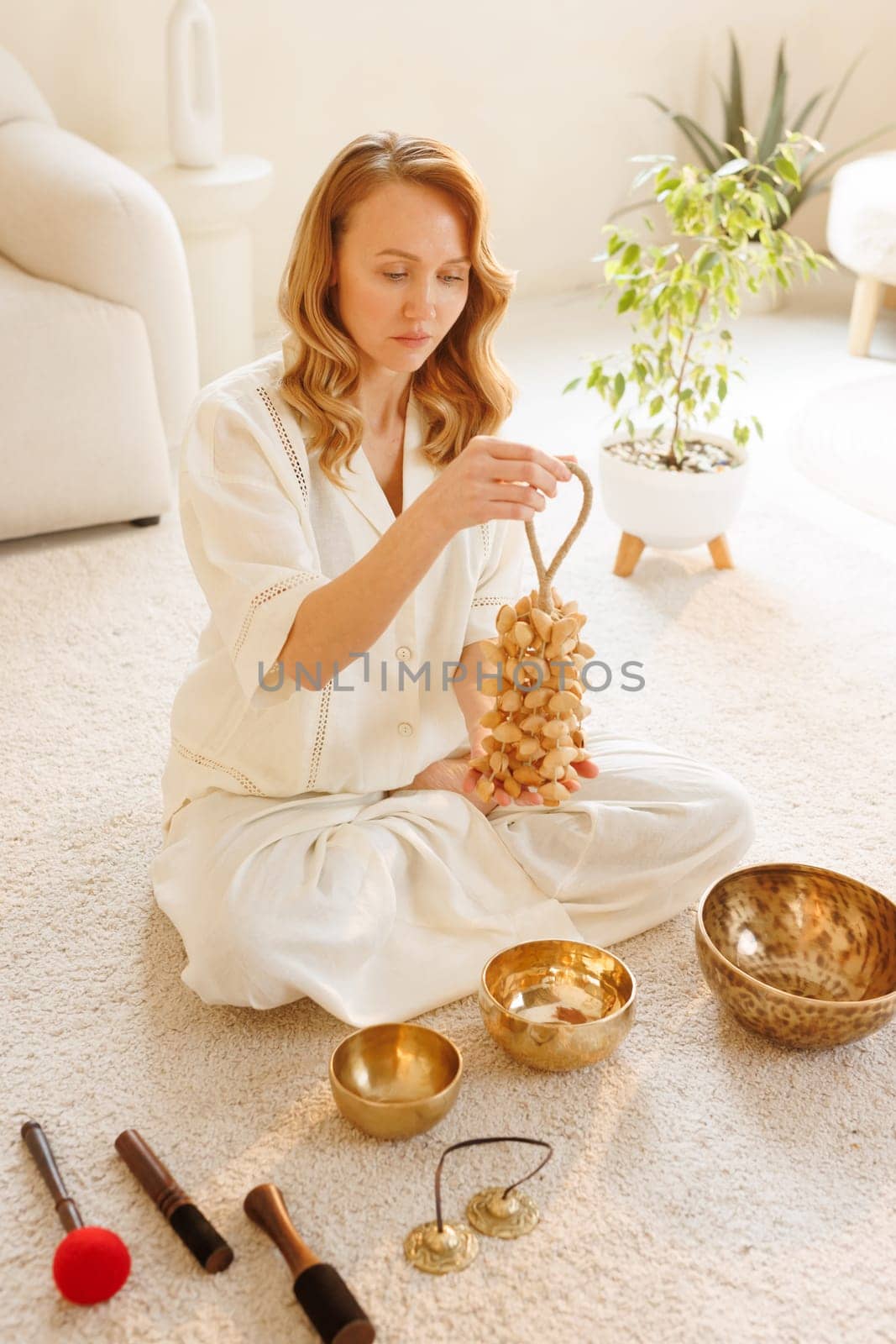 Woman in white meditating at home with spiritual instruments, holding rattle seeds for sound therapy and balance.