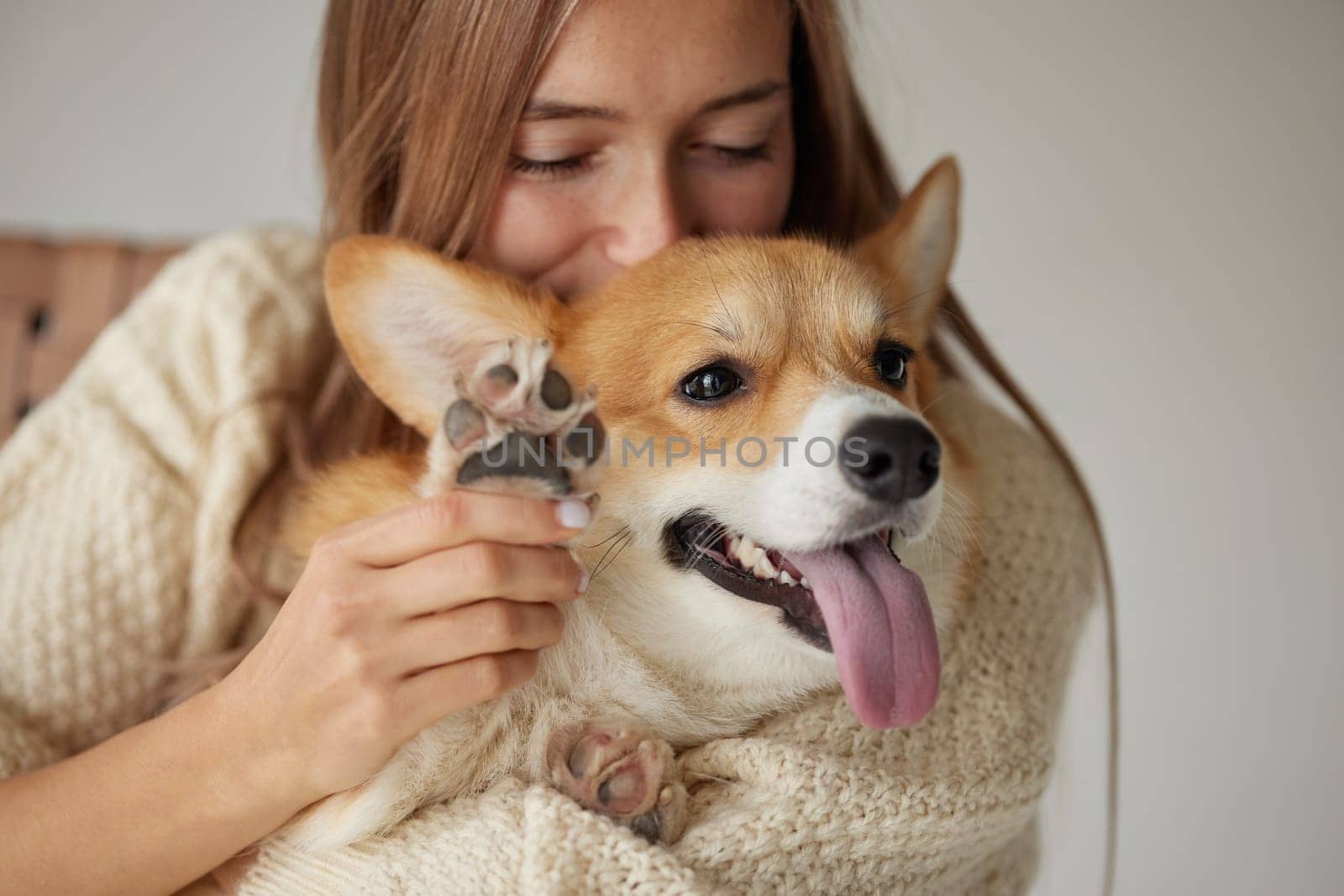 Smiling woman cuddling happy Corgi dog, holding paw up, close bonding moment showing love and affection indoors.