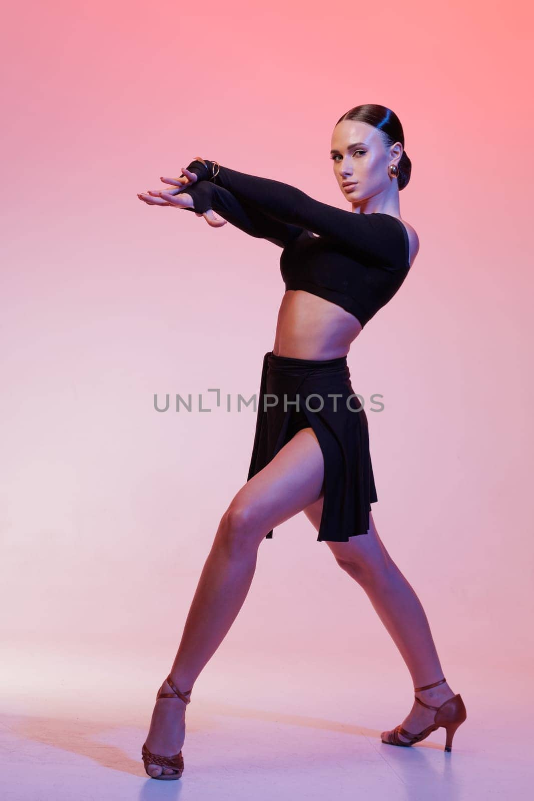Confident female Latin dancer in stylish black costume performing dramatic pose against pink gradient background.