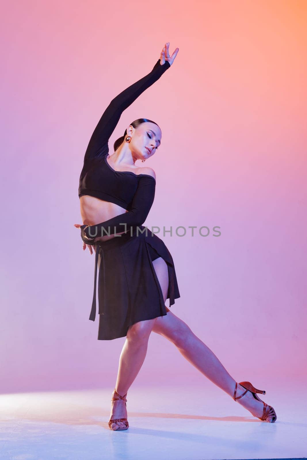 Graceful female dancer in elegant black costume striking a dramatic pose under pink-orange studio lighting.