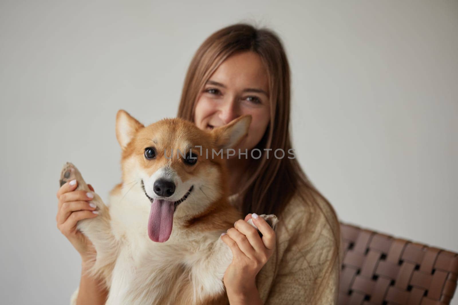 Cheerful woman holding a happy Corgi dog raising its paws, joyful friendship moment, cozy home vibe, neutral background.