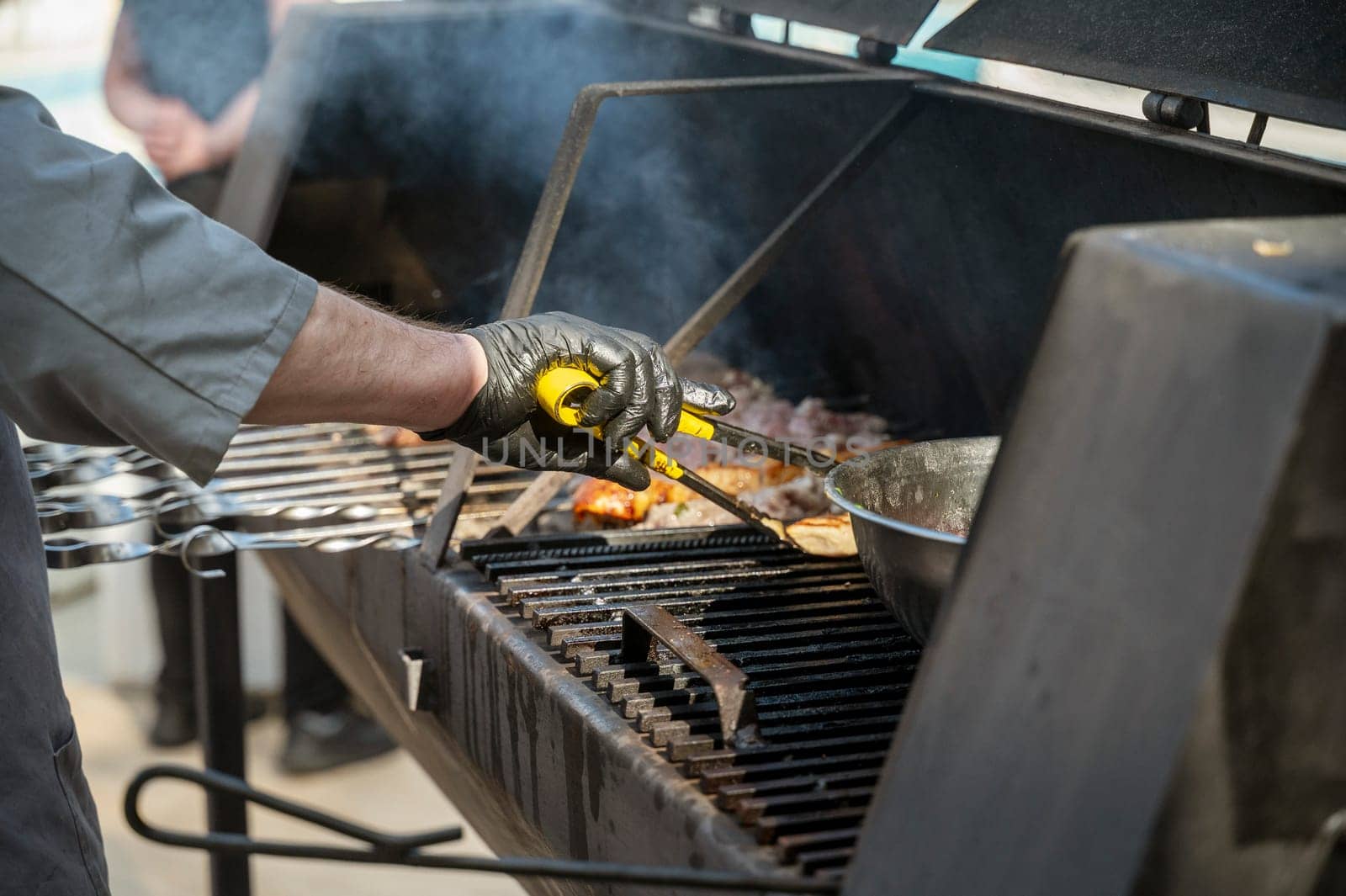 Male chef preparing meat on grill at barbecue party in summer garden, chef's hands in gloves. Concept of food, people and family time. Pork on skewers is roasted on coals in large metal grill device