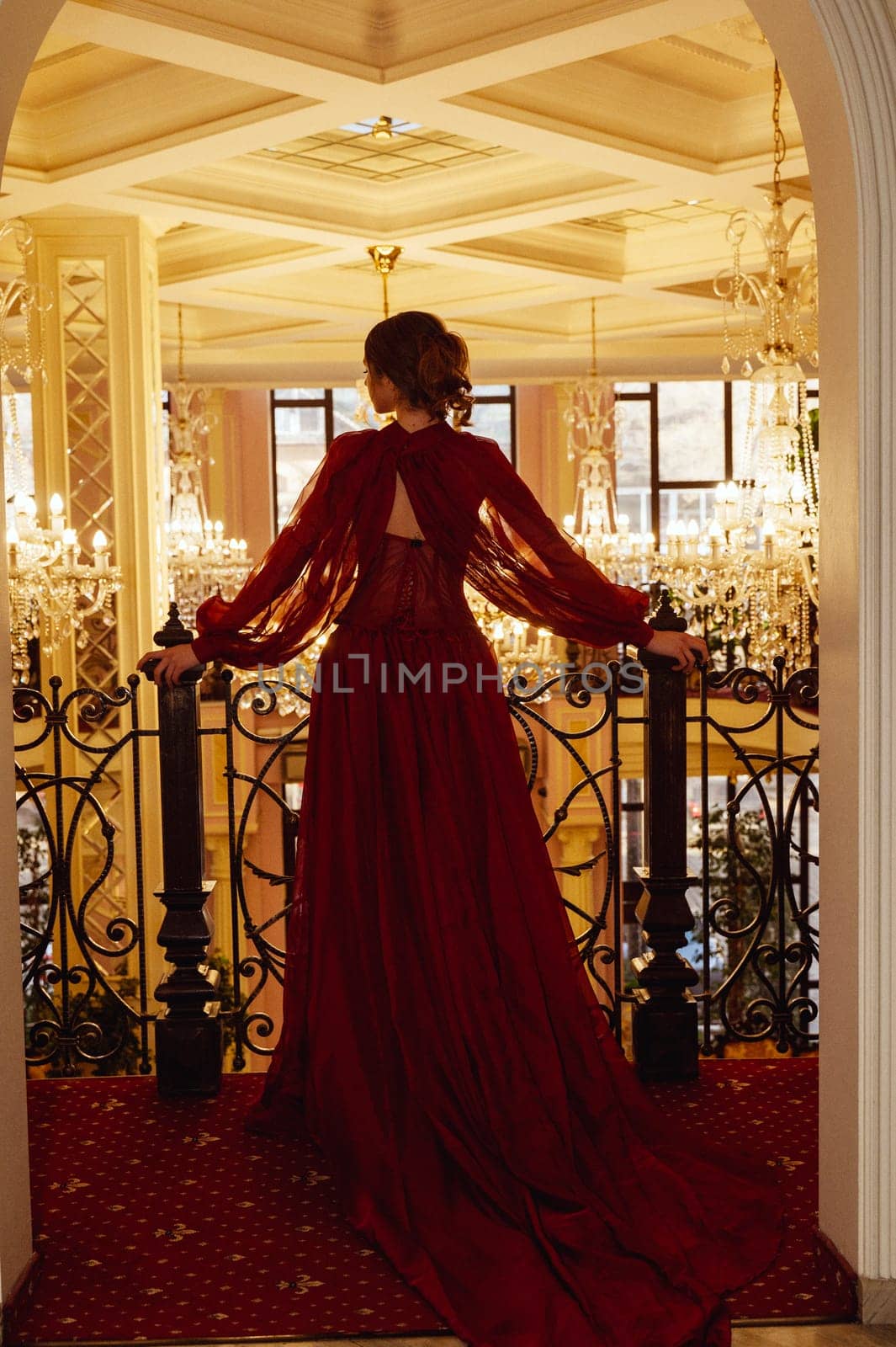 Silhouette of a model in a red dress against the backdrop of a luxurious hotel interior with crystal chandeliers. View from the back of an elegant young woman in a long evening dress