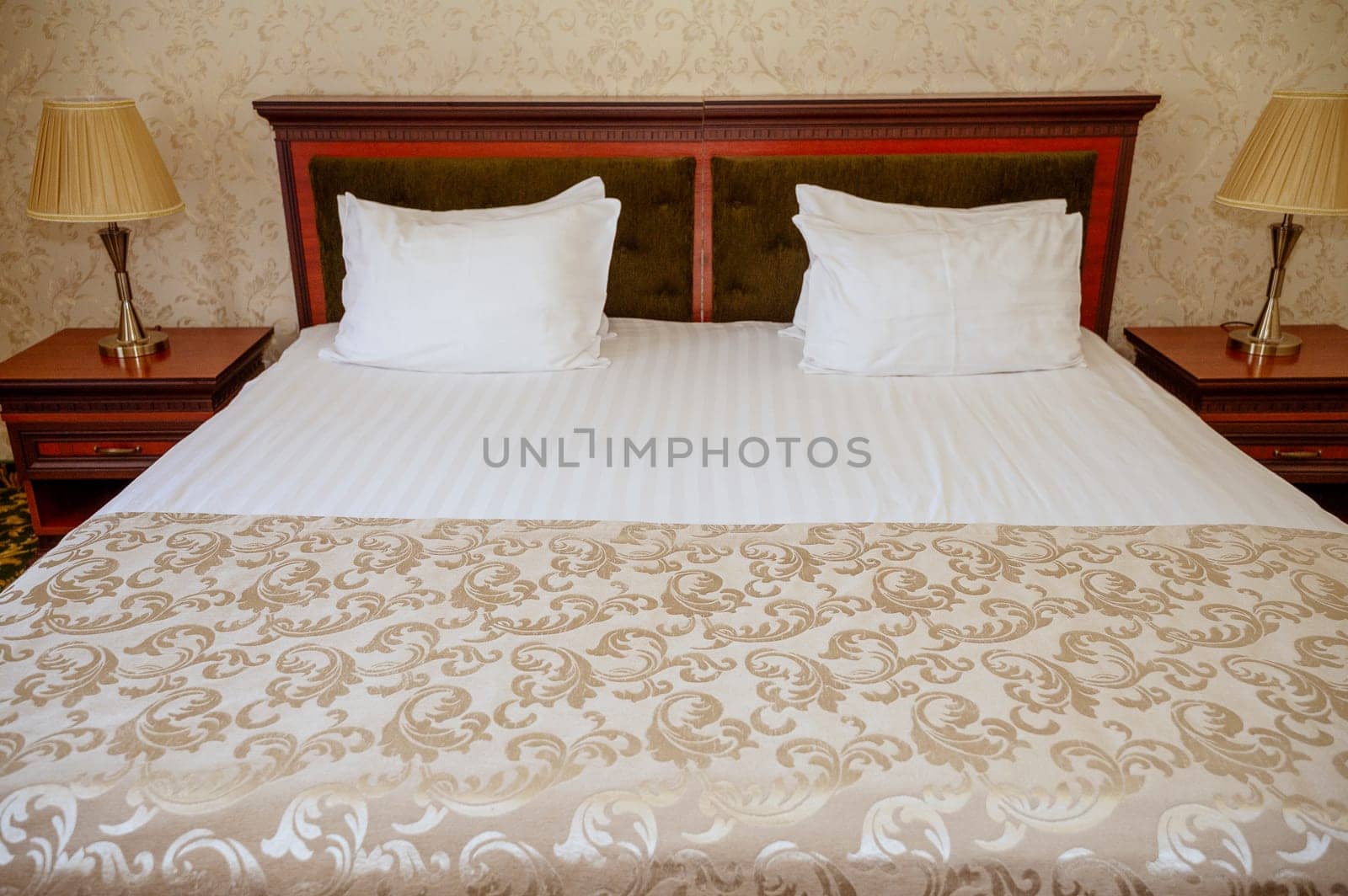 Double bed with wooden, dark red headboard, clean white bed linen and pillows. bedspread and wallpaper of the same golden beige color. On the bedside tables are table lamps with golden lampshades