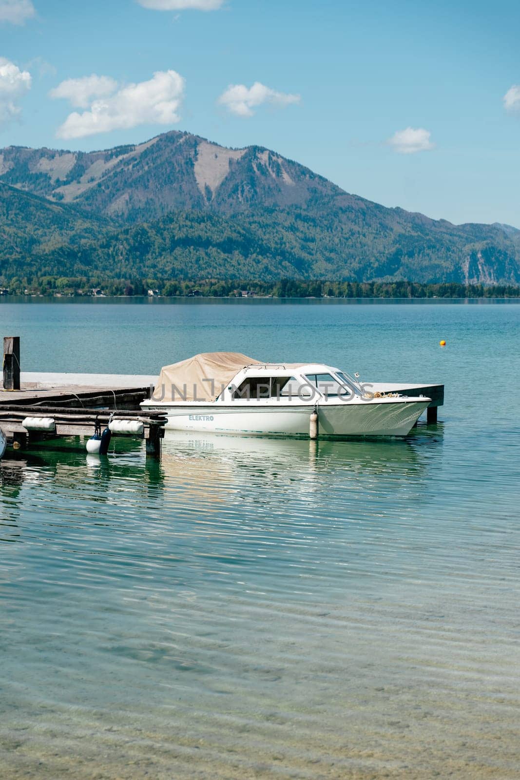 A boat rests at the dock of Wolfgangsee in Strobl, Upper Austria. Surrounded by mountains, the calm lake reflects the clear sky, capturing the essence of spring and summer relaxation.