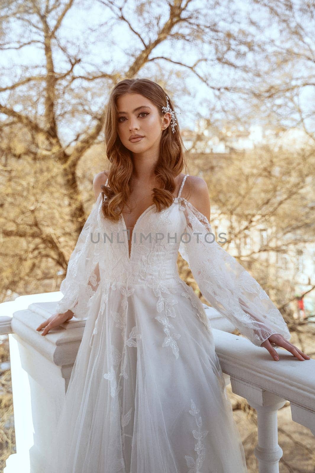 Young brunette bride in traditional long white dress posing on hotel balcony in spring. Long white dress with long sleeves, corset and open shoulders. Bride portrait, wedding makeup
